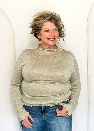 A smiling woman with short curly gray-blonde hair wearing a light beige long-sleeve knit top and blue jeans, standing against a white arched backdrop.