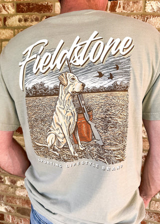 A man viewed from behind wearing a light sage green t-shirt with a large graphic print on the back. The graphic features a hunting dog holding a shotgun with a shotgun shell bag, under the text 'Fieldstone' and 'Sporting Lifestyle Brand'.