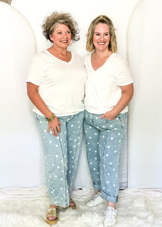 Two women standing side-by-side wearing matching white v-neck t-shirts and light blue jeans with white polka dots, posed in front of a white studio backdrop with arches.