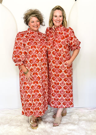Two women standing side-by-side wearing matching red and white patterned midi dresses with puff sleeves and high necks, posed in front of a white backdrop with arches.