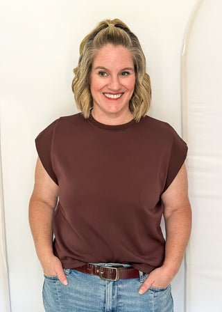 A smiling woman with blonde hair tied back stands against a white arched backdrop, wearing a sleeveless dark brown top and light blue jeans with a brown belt.