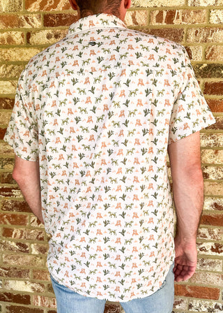 A man viewed from behind wearing a short-sleeve button-up shirt with a white base and a repeating pattern of small orange and green motifs, paired with light blue jeans, standing against a brick wall.
