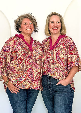 Two women standing side by side in matching paisley patterned tops and blue jeans, posing in front of a white arched backdrop in an indoor room.