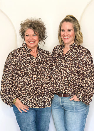 Two women standing side by side wearing matching brown and black leopard print button-down shirts and blue jeans, posing in front of a white arched backdrop.
