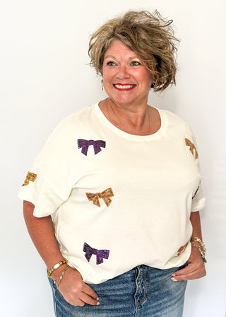 Short sleeve white top featuring scattered sequin bows in gold and purple