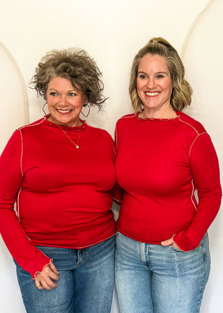 Two women standing side-by-side wearing matching bright red long-sleeve mock neck tops and blue jeans, posing against a white arched backdrop.