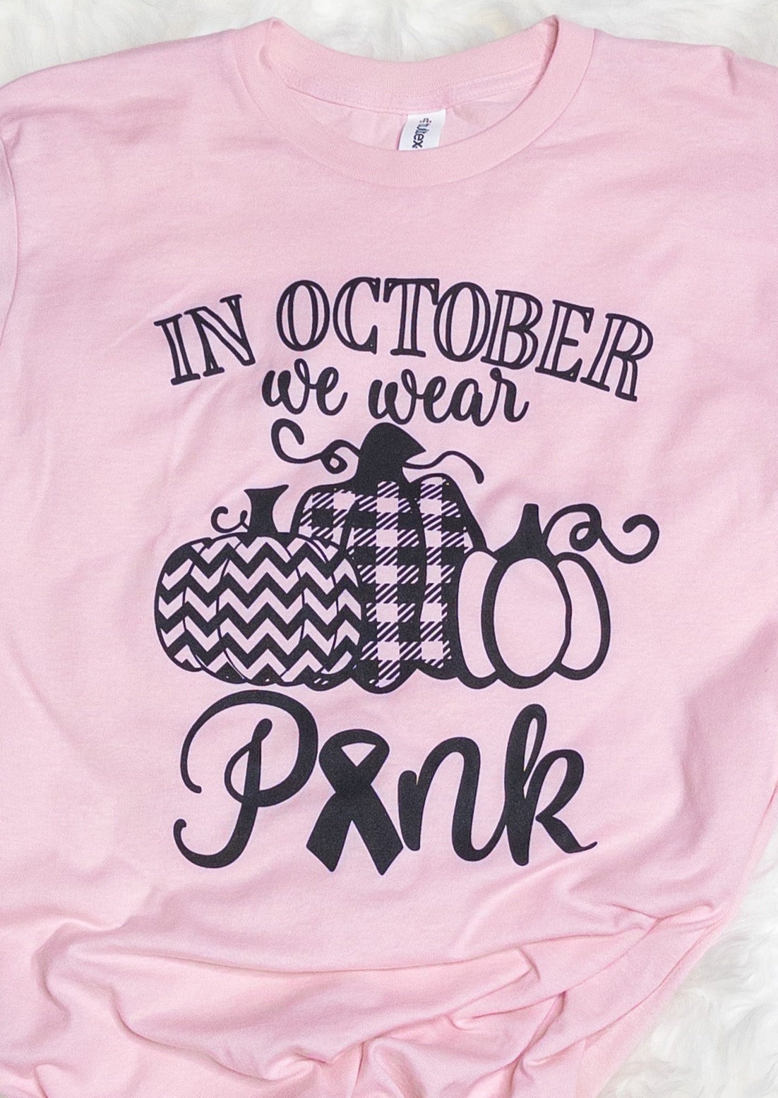 A light pink colored tee shirt that says "In October we wear pink". It has three pumpkins, each with a different pattern.