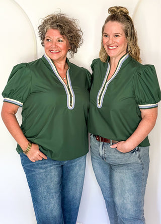 Two women standing side by side wearing matching dark green puff-sleeve tops with a V-neckline, white trim, and gold-toned zipper detail, paired with blue jeans against a white arched backdrop.