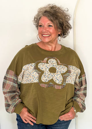 woman wearing an olive green  patchwork top with floral fabric pattern on the front and plaid long sleeves. 
