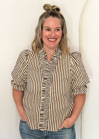 A smiling woman with blonde hair in a bun stands against a white arched wall, wearing a brown and white vertically striped short-sleeve blouse with ruffled trim and blue jeans.