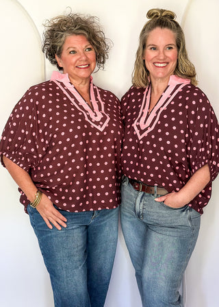Two women standing side-by-side wearing matching burgundy and pink polka dot tops with pink trim, paired with blue jeans, posing in front of a white studio backdrop.