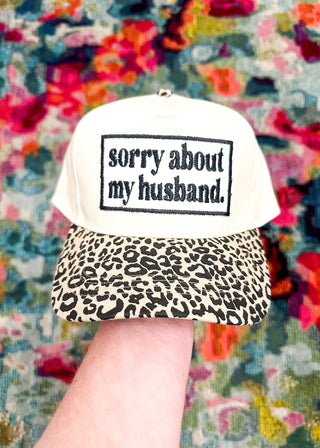 Cream snap back ball cap with a leopard bill and embroidered patch “sorry about my husband,” stitched in black.