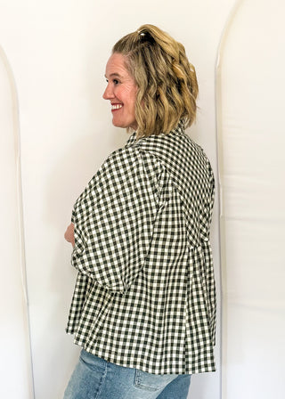 A woman with blonde hair tied back is shown from the side, wearing a green and white gingham button-up shirt and blue jeans, standing against a white arched backdrop.