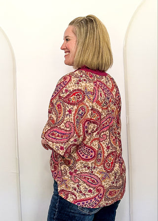 A woman with blonde hair stands in profile wearing a red and beige paisley patterned long-sleeve shirt and dark blue jeans, standing in front of three large white arch-shaped inflatable backdrops in an indoor room with a ceiling fan visible.