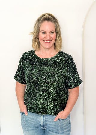 A woman with blonde hair wearing a dark green sequined short-sleeve top and light blue jeans, standing against a white arched backdrop.
