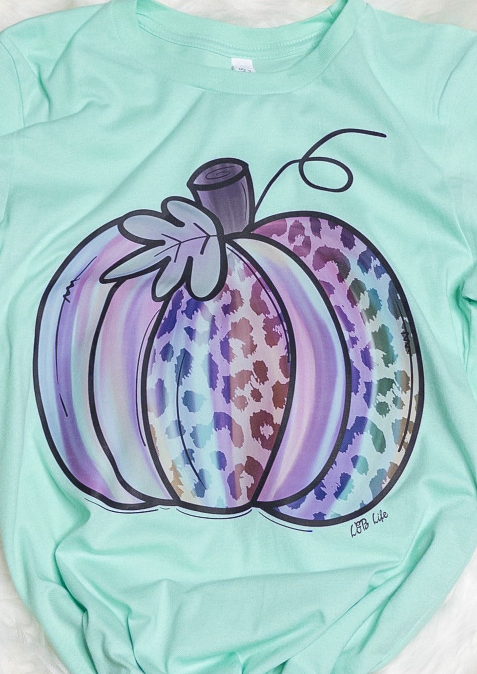 A mint colored tee shirt with a pumpkin. Pumpkin has some leopard print detailing and is light water color design with blues, purples, and pinks 