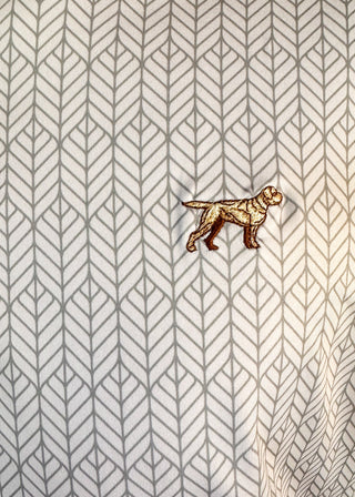 A close-up of a white fabric with a repeating gray geometric leaf pattern and a small embroidered dog motif in brown and beige near the center-right.