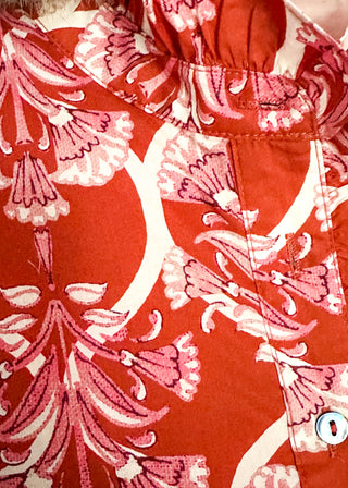 A close-up of a red-orange blouse with a pink and white floral pattern, featuring a ruffled neckline and a buttoned pocket.
