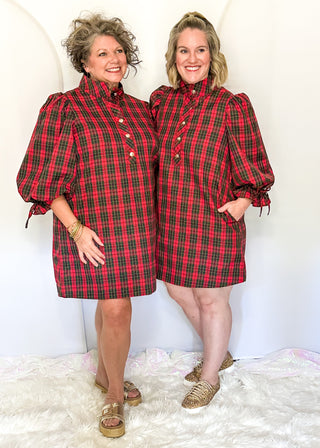 Two women modeling matching red and green plaid puff-sleeve mini dresses with high mock necks and front buttons, standing on a white faux fur rug against a white studio backdrop.