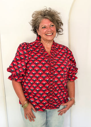 A smiling woman with curly gray hair wearing a red and black patterned blouse with light blue jeans, standing against a white arched backdrop.