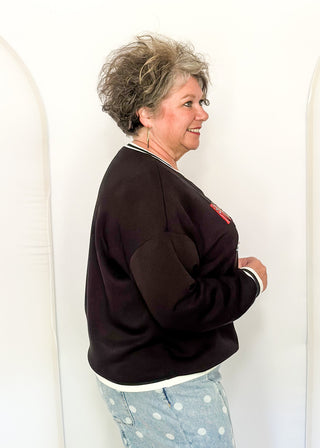 A woman with short gray hair is shown from the side, wearing a black crewneck sweatshirt with white striped trim and a small red graphic on the chest, paired with light blue polka dot pants, set against a white arched studio backdrop.