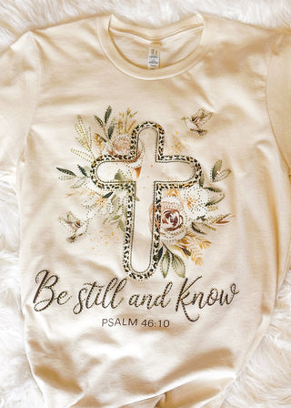 Neutral graphic tee with cross design, “Be still and know” scripture, and flat rhinestone accents