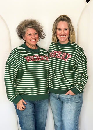 Two women standing side-by-side wearing matching green and white striped crewneck sweaters with 'MERRY' across the chest, paired with blue jeans, in front of a white arched backdrop.