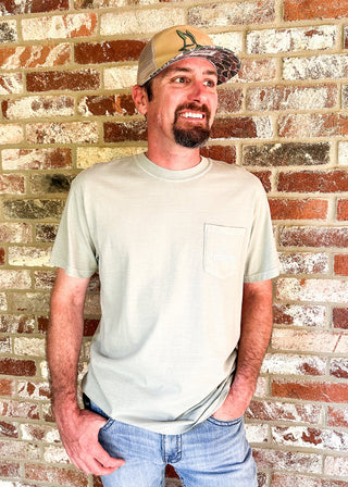 A man wearing a light sage green pocket t-shirt and a camouflage baseball cap stands against a rustic red and white brick wall.