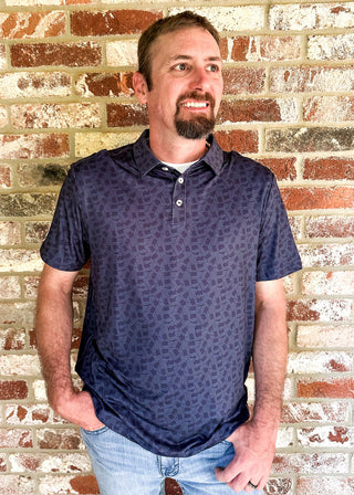 A man wearing a dark navy blue patterned short-sleeve polo shirt and light blue jeans, standing against a red and white brick wall.