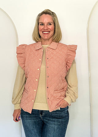 A smiling blonde woman stands indoors between large white arched panels, wearing a peach-and-white gingham ruffled sleeveless vest over a beige long-sleeve top with blue jeans.