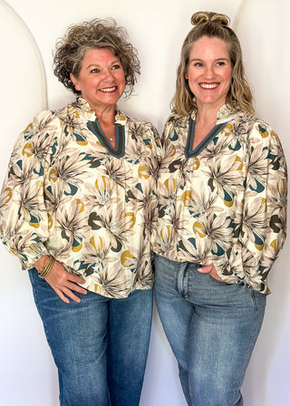 Two women standing side-by-side wearing matching floral long-sleeve collared shirts and blue jeans, posing against a white arched studio backdrop.