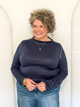 A smiling woman with curly gray hair wearing a dark navy long-sleeve top and blue jeans, standing against a white arched backdrop.