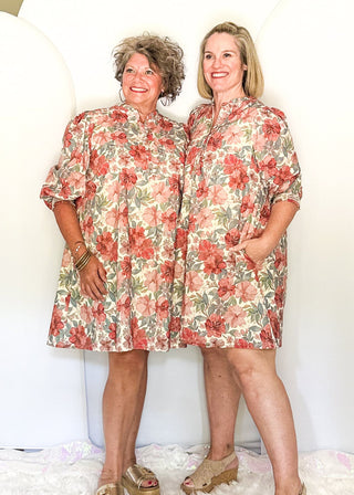 Two women wearing matching floral short-sleeve dresses stand in front of a large white photo backdrop in a home interior with hardwood floors and a ceiling fan visible.