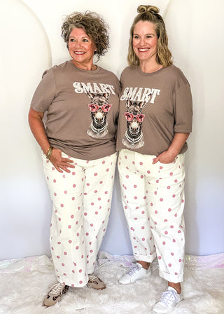 Two women standing side-by-side wearing matching outfits consisting of a brown graphic t-shirt with 'SMART' and a dog wearing sunglasses, paired with white pants featuring pink polka dots.