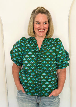A smiling blonde woman wearing a green short-sleeve blouse with a repeating teal and black scallop pattern, paired with light-wash jeans, standing against a white arched wall.