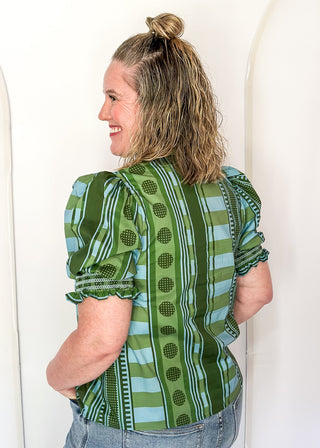 A woman shown from the back wearing a green and light blue patterned blouse with puff sleeves and scalloped edges, paired with light-wash jeans, standing against a white arched wall.