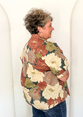 A woman with short curly gray hair is shown from the back and side, wearing a short-sleeve button-up shirt with a large floral print in rust red, cream, and olive green, paired with blue jeans, standing against a white arched wall.