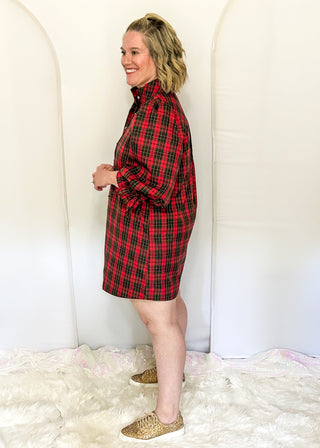 A woman modeling a red and black plaid shirt dress, standing in front of a white studio backdrop with arches and a white faux fur rug.