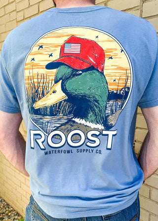 dusty blue tee shirt with duck head in the slew and red hat on with american flag - roost waterfowl supply co on the back - front pocket
