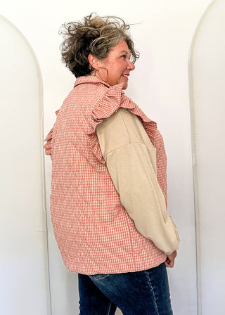 A woman wearing a pink and white gingham quilted vest with beige ruffled shoulder details and long beige sleeves, paired with blue jeans, standing in front of white arched panels.