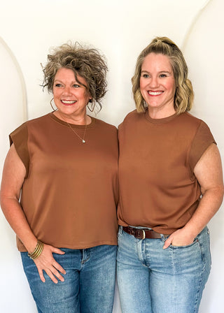 Two women standing side-by-side wearing matching brown short-sleeve crewneck tops and blue jeans, posing in front of a white arched backdrop.