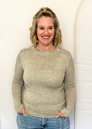 A smiling blonde woman wearing a light beige long-sleeve knit sweater and blue jeans, standing against a white arched backdrop.