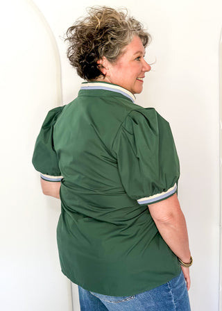 A woman wearing a dark green short-sleeve blouse with puffed sleeves and striped trim on the collar and cuffs, paired with blue jeans, standing against a white arched wall.