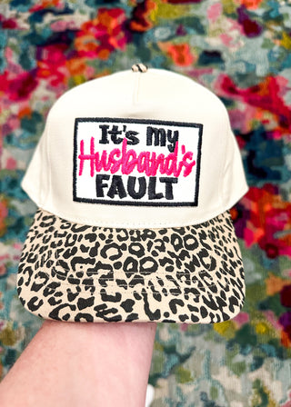 Cream snap back ball cap with a leopard bill and embroidered patch “It’s my Husbands Fault,” stitched in hot pink and black.