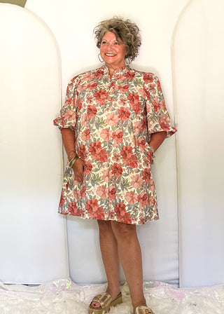 A woman stands in front of three large white arch backdrops in a home studio setup, wearing a short-sleeve floral dress with red, pink, and green tones.