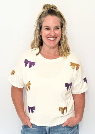 Short sleeve white top featuring scattered sequin bows in gold and purple