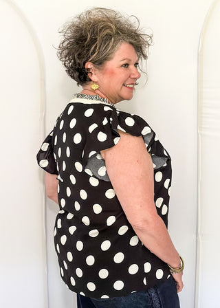 Women's black blouse with cream polka dots, striped split neckline, and flutter sleeves.