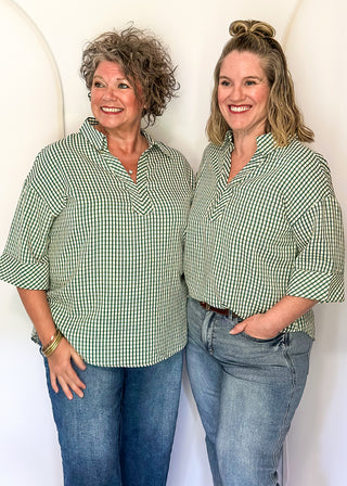 Two women standing side-by-side wearing matching green and white gingham button-down shirts with blue jeans, posing against a white studio backdrop with arched shapes.