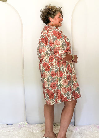 A woman wearing a red and green floral print short-sleeve dress stands in front of three large white arched photo backdrops in a home setting with a ceiling fan visible.