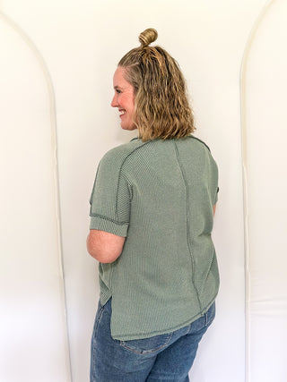 Woman wearing a green patterned shirt and blue jeans standing against a white wall.